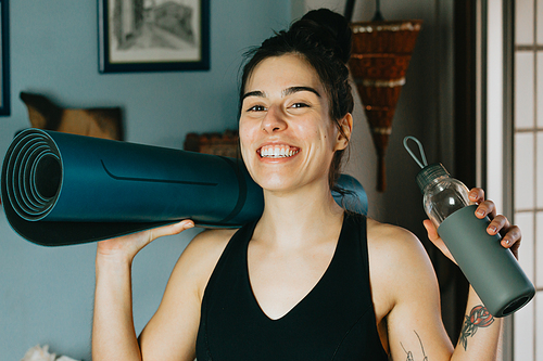 Young woman smiling to camera while holding a yoga mat and a bottle of water at a modern and bright flat