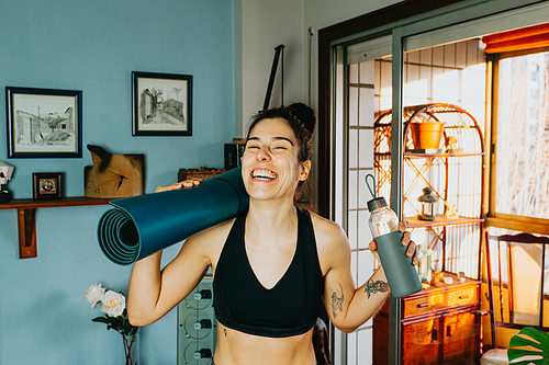 Young woman smiling and laughing to camera while holding a bottle of water and a yoga mat at a modern flat in the city during a bright day