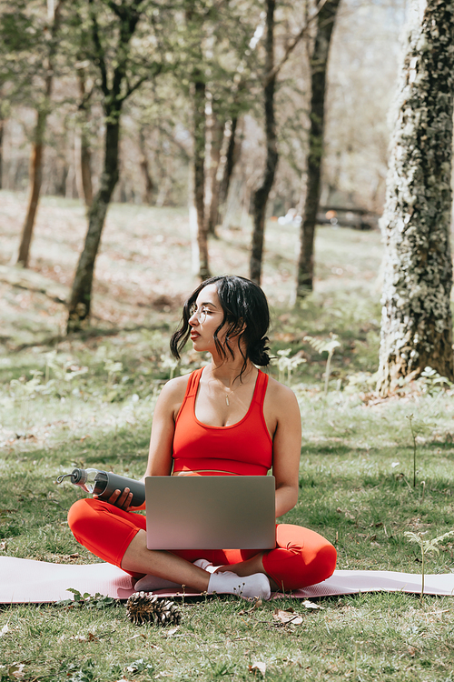 Young woman in fitness clothes relaxing on his laptop grabbing a bottle of water at the park over a yoga mat