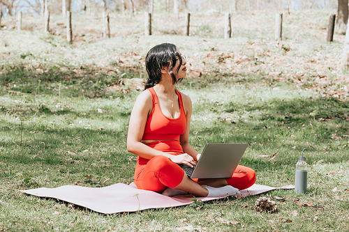 Young woman in fitness clothes relaxing on his laptop at the park over over a yoga mat with copy space