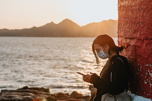 Young woman using his phone at the coast of spain with a massive sunset as the background while using a mask relax concept