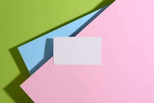 blank rectangular business card lies on a modern bright green background with blue and pink sheets of paper with a shadow. Business template, flat lay