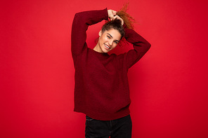 Young beautiful curly brunette woman with sincere emotions poising isolated on background wall with copy space wearing casual dark red sweater. Fun and joy concept.