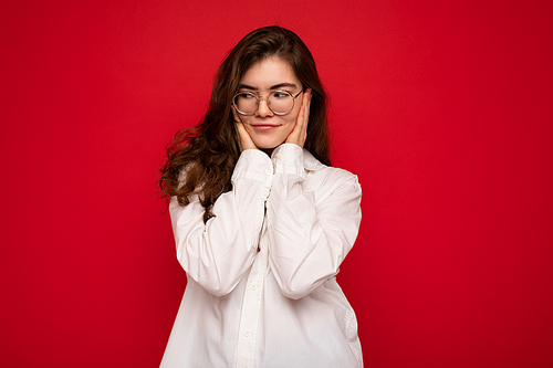 Attractive young curly brunette woman wearing white shirt and optical glasses isolated on red background with empty space.
