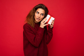 Photo shot of attractive serious thoughtful concentrated young brunette curly woman isolated over red background wall wearing red sweater holding gift box looking to the side and thinking.