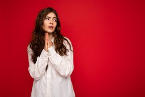 Portrait of beautiful young curly brunette woman wearing white shirt isolated on red background with copy space, praying and dreaming with sincere emotions.