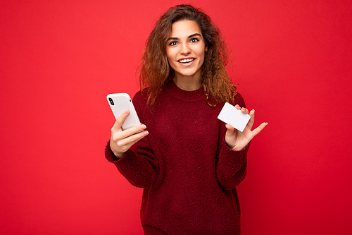 Young delightful attractive brunette female person with sincere emotions isolated over background wall with empty space wearing casual dark red sweater holding credit card and using mobile phone . Bank concept.