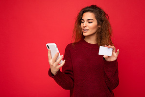 Young beautiful curly brunette woman with sincere emotions poising isolated over background wall with empty space wearing casual dark red sweater holding credit card and using mobile phone . Internet concept.