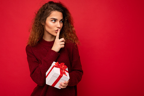 Photo shot of beautiful positive young brunette curly woman isolated over red background wall wearing red sweater holding gift box looking to the side and showing shh gesture. Copy space