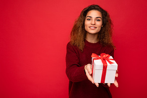 Beautiful happy young brunette woman isolated over colourful background wall wearing stylish casual clothes holding gift box and looking at camera.