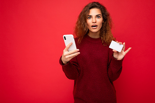 Young beautiful curly brunette woman with sincere emotions poising isolated over background wall with empty space wearing casual dark red sweater holding credit card and using mobile phone . Shock and surprise concept.