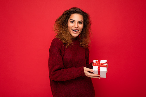 Attractive positive surprised young brunette curly woman isolated over red background wall wearing red sweater holding gift box looking at camera.