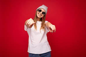Portrait of young emotional positive happy smiling beautiful dark blonde woman with sincere emotions wearing casual white t-shirt with empty space for mockup gray hat and sunglasses isolated over red background with free space and pointing at camera.