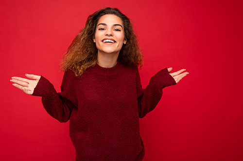 Photo of young positive happy beautiful brunette curly woman with sincere emotions wearing casual red pullover isolated on red background with copy space.