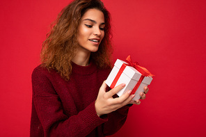Shot of attractive positive smiling young brunette curly woman isolated over red background wall wearing red sweater holding gift box looking at white box with red ribbon and thinking.
