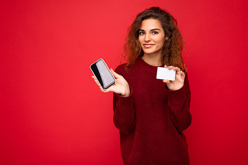 Young beautiful curly brunette woman with sincere emotions poising isolated over background wall with empty space wearing casual dark red sweater holding credit card mobile phone with empty screen display for mock up. online concept.