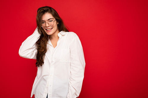 Portrait of beautiful positive cheerful cute smiling young brunette woman in casual white shirt and trendy optical glasses isolated on red background with copy space.