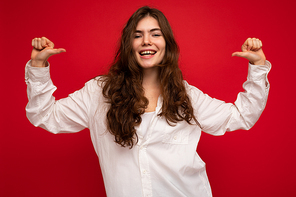 Photo of young beautiful happy smiling curly brunette woman wearing white shirt. Sexy carefree female person posing isolated near red wall in studio with free space. Positive model with natural makeup.