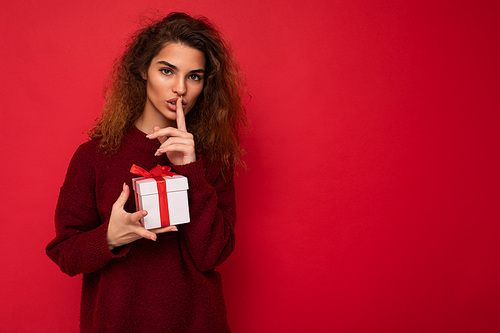 Shot of pretty young brunette curly woman isolated over red background wall wearing red sweater holding gift box looking at camera and showing shh gesture and meaning be quiet. Copy space, mockup