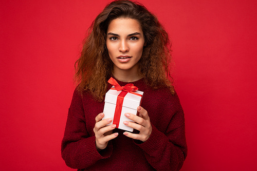Charming self-confident young brunette curly woman standing isolated over red background wall wearing dark red sweater holding gift box looking at camera.