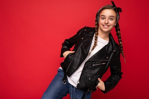 Photo shot of beautiful happy smiling brunette little girl with pigtails wearing trendy black leather jacket and white t-shirt standing isolated over red background wall looking at camera. Copy space