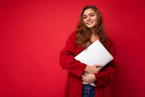 Close-up portrait of Beautiful smiling young brunette woman holding netbook computer looking at camera wearing red cardigan isolated on red wall.