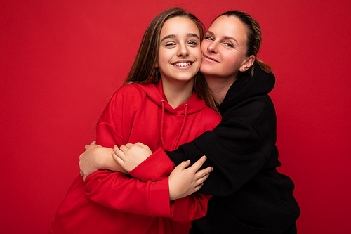 Photo of beautiful happy smiling young daughter wearing stylish red hoodie and adult mother wearing trendy black hoodie isolated over red background looking at camera and having fun.