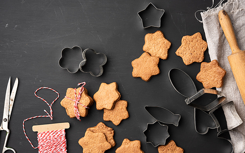 baked star shaped gingerbread cookies, wooden rolling pin and metal cutters on a black table, top view