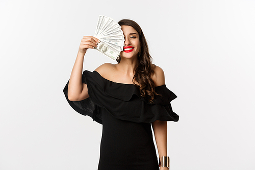 Fashion and shopping concept. Happy young woman in black dress, with red lips, holding money and smiling satisfied, standing over white background.