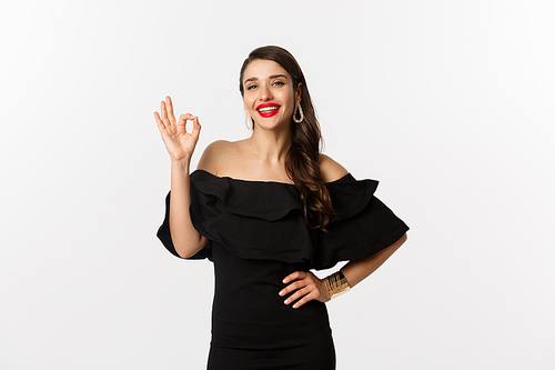 Fashion and beauty. Satisfied good-looking woman with red lipstick, black dress, showing okay sign in approval, like and agree, standing over white background.