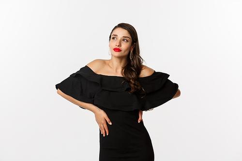 Fashion and beauty. Stylish modern woman in black dress, makeup and red lips, posing over white background self-assured, standing over white background.