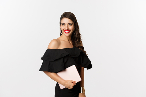 Fashion and shopping concept. Stylish young woman with glamour makeup, wearing black dress, holding digital tablet and smiling, white background.