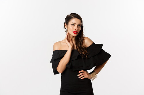 Sensual young woman in black dress, showing her earrings and looking sexy at camera, standing over white background.