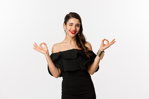 Fashion and beauty. Attractive brunette woman in black dress, showing okay signs and winking at camera, approve and recommend, standing over white background.