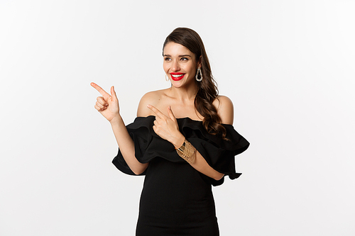Fashion and beauty. Attractive woman in jewelry, makeup and black dress, laughing and pointing fingers left at promo offer, white background.