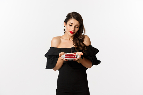 Party and celebration. Cute young woman with red lips, wearing black dress, holding birthday cake with candle and looking dreamy, making wish, white background.