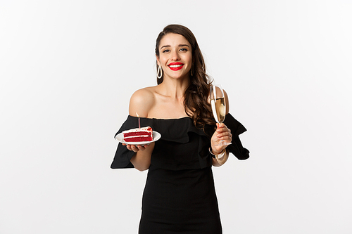 Celebration and party concept. Happy woman holding birthday cake and drinking champagne, smiling while standing in black dress against white background.