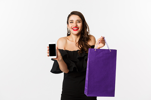 Beauty and shopping concept. Beautiful and stylish woman showing mobile phone screen and bag, buying online, standing over white background.