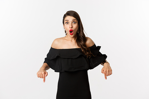 Fashion and beauty. Elegant woman in black dress pointing fingers down, showing promo and smiling, standing over white background.