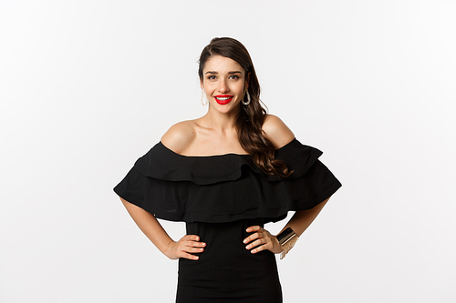 Waist-up shot of beautiful woman with red lipstick, wearing black dress and smiling pleased, standing over white background.