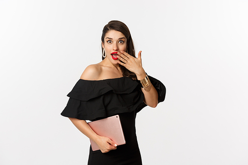Fashion and shopping concept. Stylish young woman with glamour makeup, wearing black dress, holding digital tablet and looking surprised, white background.