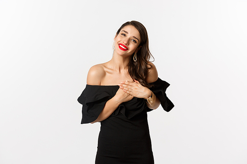 Fashion and beauty. Attractive glamour woman in black dress saying thank you, smiling and holding hands on heart with pleased emotion, white background.