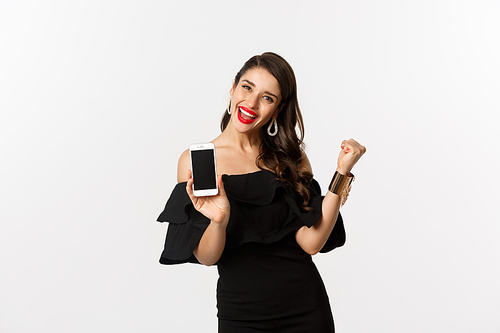 Online shopping concept. Satisfied pretty woman showing smartphone screen, making fist pump to rejoice, winning in internet, standing over white background.