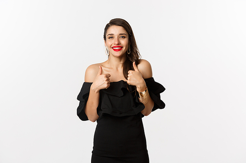 Fashion and beauty. Gorgeous woman in black dress, red lipstick, showing thumbs up in approval, recommending product, standing over white background.
