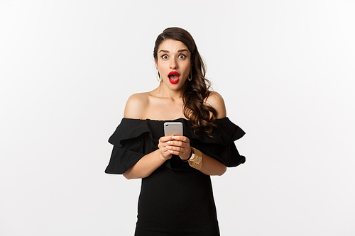 Happy young glamour woman in black dress, holding mobile phone and looking surprised, standing over white background.
