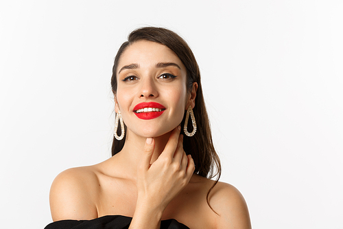 Fashion and beauty concept. Close-up of gorgeous brunette woman with red lips, touching face and smiling self-assured, standing over white background.