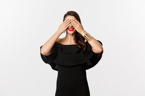 Fashion and beauty. Beautiful woman in black dress and red lipstick, covering eyes and smiling, waiting for surprise, standing over white background.
