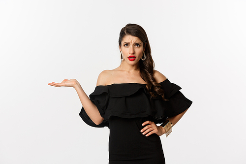 Fashion and beauty. Annoyed woman in black dress raising hand, so what gesture, looking confused at camera, standing bothered over white background.