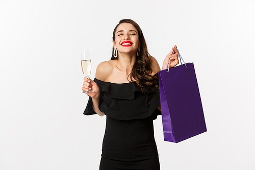 Happy smiling woman celebrating, holding present in shopping bag and glass of champagne, standing in black dress over white background.