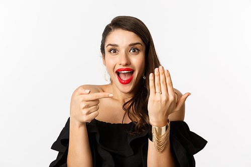 Happy young woman saying yes, become a bride, pointing at engagement ring on finger and smiling pleased, standing over white background.